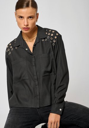 Chemise noire avec des accents de œillets argentés sur les épaules, deux poches avant, design à boutons, et manches longues avec des poignets à bouton.
