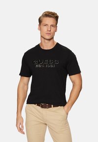 Camiseta negra de manga corta hecha de algodón, con un logotipo en relieve "GUESS EST. 1981" en un acabado brillante en el pecho.