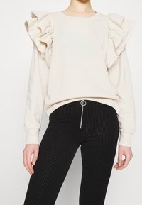 Beige sweatshirt med stora volangärmar, rund halsringning och en avslappnad passform, tillsammans med svarta ribbade kordbyxor med dragkedjedetalj.