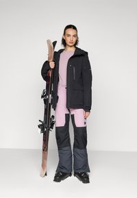 Slēpošanas apģērbs, kas ietver melnu ūdensizturīgu jacket, gaiši rozā topu un rozā-melnas slēpošanas bikses. Turēšana slēpes ar melniem stiprinājumiem, elegants dizains.