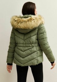 Vrouw in een olijfgroene gewatteerde jas met capuchon met bontvoering, met de rug naar de camera staand tegen een effen lichte muur.