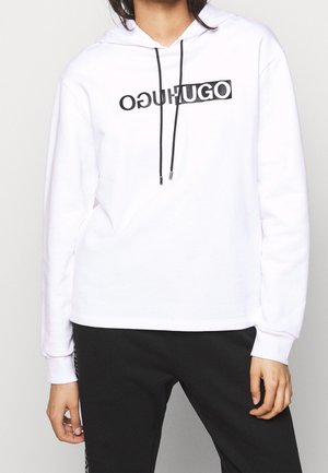 Person iført en hvid hættetrøje med sorte snører og et spejlvendt "HUGO" logo på brystet, kombineret med sorte bukser.