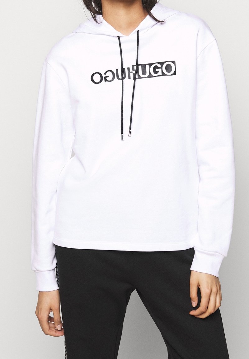 Personne portant un sweat à capuche blanc avec des cordons noirs et un logo "HUGO" en miroir sur la poitrine, assorti d'un pantalon noir.