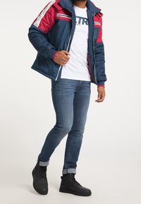 Veste matelassée bleu marine et rouge avec une fermeture éclair intégrale, des poignets côtelés et deux poches latérales ; associée à un jean bleu et des bottes noires à lacets.