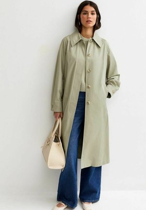 Vrouw draagt een lichtgroene trenchcoat, blauwe jeans met wijde pijpen, beige trui, witte schoenen, en draagt een beige handtas, staand tegen een witte muur.