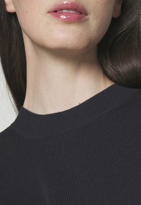 Granatowy sweter prążkowany z wysokim dekoltem, teksturowana tkanina i subtelne detale przeszyć widoczne przy kołnierzu. Gładka skóra i błyszczące usta.