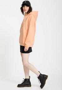 Jasnopomarańczowy hoodie z przednią kieszenią, zestawiony z czarną mini spódnicą i czarnymi wiązanymi botkami. Czapka z logo z przodu.