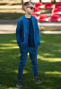 Niebieski sportowy dres z zapinanymi na zamek kurtką i dopasowanymi spodniami, wykonany z gładkiego materiału. Noszony z ciemnymi okularami przeciwsłonecznymi i zielonymi trampkami.