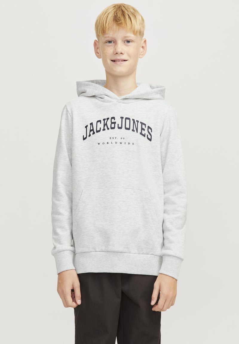 Blond chłopiec w jasnoszarej bluzie z kapturem z napisem "JACK&JONES" i czarnych spodniach, stojący na jednolitym jasnym tle.