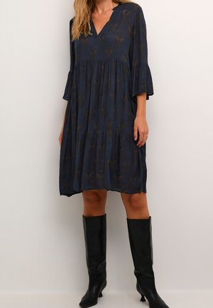 Femme portant une robe ample bleu marine avec des motifs subtils, manches trois-quarts et bottes noires à talons montant jusqu'au genou, debout devant un fond uni.