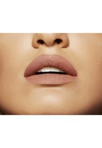 Rouge à lèvres nude mat sur des lèvres pleines avec une texture lisse. Subtil éclat sur les dents, fond de teint naturel.