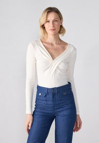 Langermet, kremfarget bodysuit med dyp v-hals og vridt detalj foran, kombinert med høyt liv, mørkeblå denimjeans med lapper.