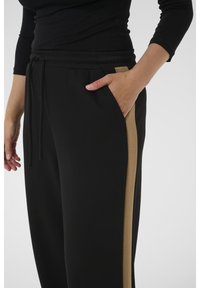 Oseba, ki nosi črne hlače z bež stranskimi črtami in elastičnim pasom, z roko naslonjeno v desnem žepu na belem ozadju.