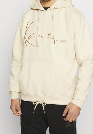 Sudadera color crema con un bolsillo frontal, capucha ajustable con cordón y un logo en cursiva de color marrón. Tejido suave, con puños y dobladillo acanalados.