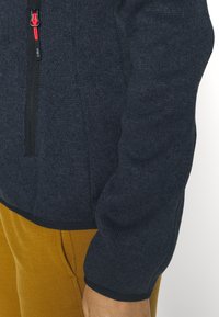 Tummansininen fleecetakki, jossa on vetoketjullinen etuosa, teksturoidusta kankaasta valmistettu ja mustalla somistuksella helman ja hihansuiden ympärillä.