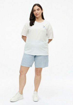 Frau steht mit den Händen hinter dem Rücken, trägt ein weißes T-Shirt mit kleinem Palmenmotiv, blaue gestreifte Shorts und weiße Sneakers.