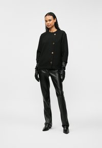 Μαύρη φόρμα με χρυσά κουμπιά, μακριά μανίκια, σε συνδυασμό με εφαρμοστά μαύρα δερμάτινα παντελόνια και ταιριαστά γάντια, αναδεικνύοντας μια κομψή, σύγχρονη εμφάνιση.