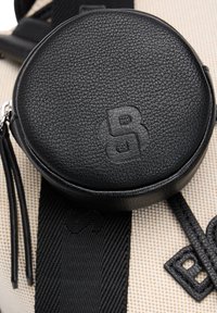 Sort læder rund taske med tekstureret finish, præget logo, fastgjort til en beige taske med sorte remme og detaljer.