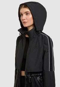 Žena oblečená v čiernej kapucňovej bunde so zipsom vpredu, reflexnými pruhmi na rukávoch a nášivkou s logom na hrudi, otočená doľava.