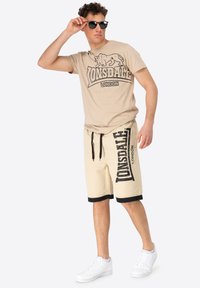 Beige Lonsdale T-shirt with logo, beige shorts with side branding, black accents, and white sneakers; sunglasses worn on the face.