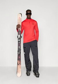 Piros hosszú ujjú sportfelső, fekete sí nadrág, bézs és rózsaszín síléceket tart fekete kötésekkel, és fekete napszemüveget visel.