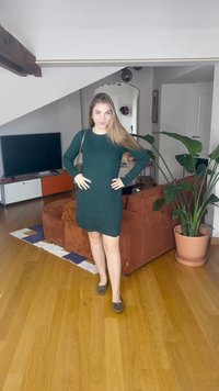 Vestido verde-escuro de malha canelada com mangas compridas, corte ajustado e decote redondo. Par de sapatos rasos castanhos. Ambiente interior com plantas.