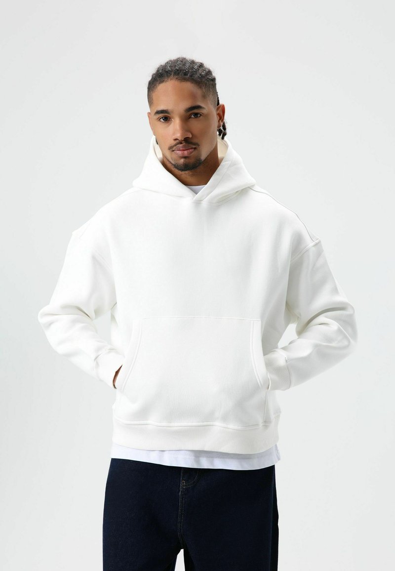 Witte hoodie gemaakt van zacht materiaal, met een kangoeroezak en geribbelde manchetten, gecombineerd met donkerblauwe spijkerbroek.