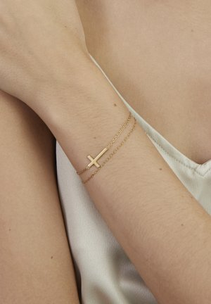 Arm wearing thin double gold chain bracelet with horizontal cross pendant over beige satin fabric garment.