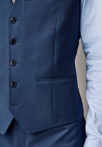 Close-up of a person wearing a blue suit vest with buttons and a light blue dress shirt sleeve visible on the right side.