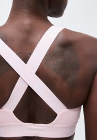 Άνω μέρος πλάτης ατόμου με λωρίδες αθλητικού σουτιέν σε ανοιχτό ροζ χρώμα που διασταυρώνονται και μια αχνή τατουάζ σε σχήμα φτερού σε σκούρο δέρμα.