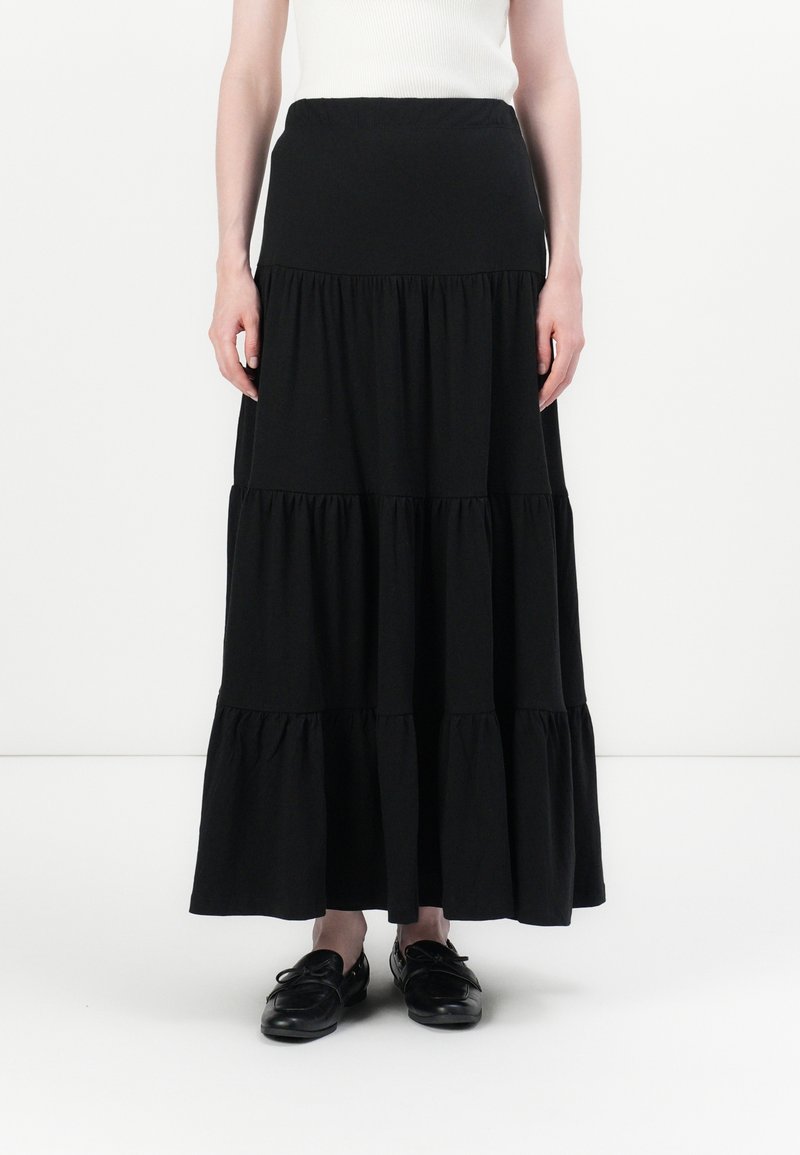 Femme portant une longue jupe noire à volants, un top blanc sans manches et des mocassins noirs, debout devant un fond blanc.