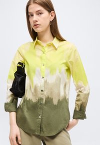 Fiatal nő hosszú ujjú gombos ingben, mely sárga, fehér és zöld átmenetes színű, fekete válltáskával, fehér háttér előtt.