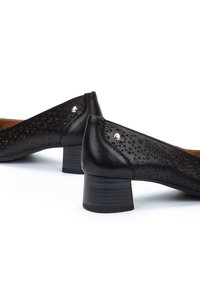 Chaussures en cuir noir avec un design perforé à l'arrière, dotées d'un petit talon carré et d'une accent métallique près du talon.