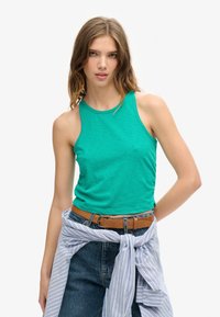 Groene mouwloze tanktop met een ronde halslijn, gecombineerd met hoge blauwe denim jeans en een lichtblauw gestreept shirt dat om de taille is geknoopt.
