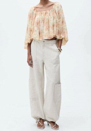 Kobieta ubrana w luźne beżowe spodnie typu cargo z elastycznymi mankietami, przezroczystą kwiatową bluzkę w kolorze brzoskwiniowym z rozkloszowanymi rękawami oraz białe sandały na paski.