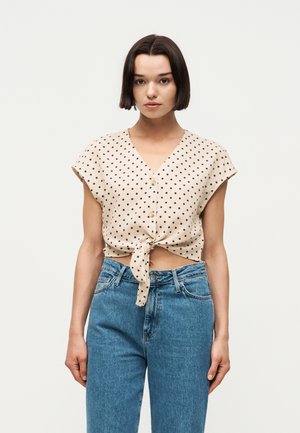 Γυναίκα φοράει μπεζ, κομμένο τοπ με κοντά μανίκια και κουμπιά, με μαύρα πουά, δεμένο στη μέση και ψηλόμεσο μπλε τζιν.