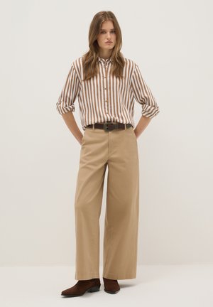 Joven de pie con las manos detrás de la espalda, vistiendo una camisa de rayas verticales marrón y blanca, pantalones beige de pierna ancha, cinturón negro y botas marrones.