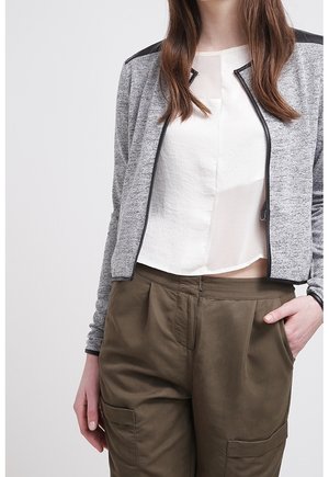 Nainen, joka pukeutunut harmaaseen vetoketjulla varustettuun takkiin mustilla yksityiskohdilla, valkoiseen puseroon ja oliivinvihreisiin korkeavyötäröisiin housuihin, joissa on taskut. Hän seisoo yhdellä kädellä taskussa.