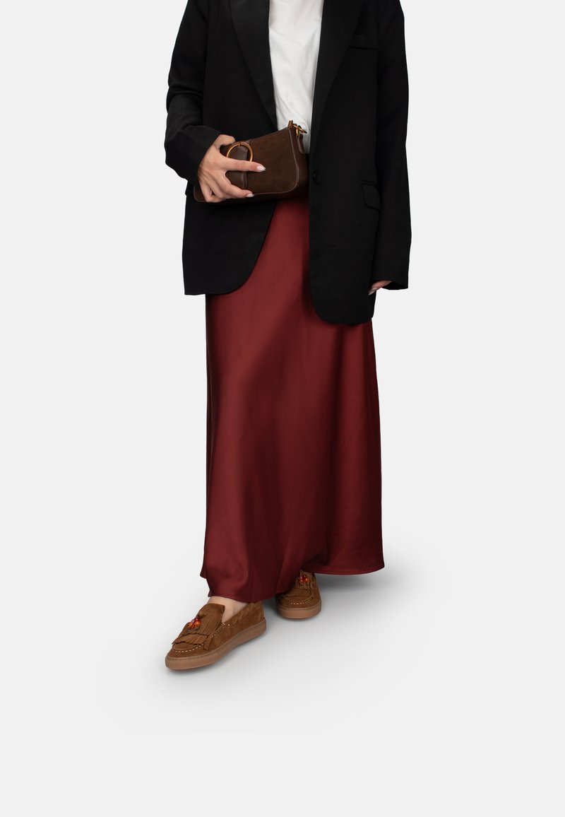 Blazer negro, camiseta blanca, falda maxi de satén burdeos, zapatos de ante marrón con borlas, sosteniendo un pequeño bolso marrón con un acento circular.