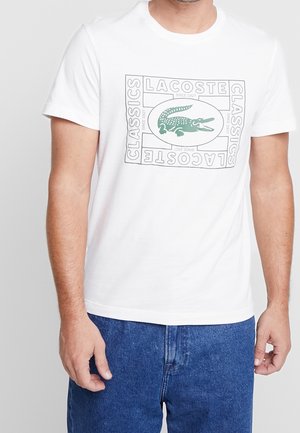 Homme portant un t-shirt blanc Lacoste avec le logo crocodile vert et le texte "CLASSICS LACOSTE DEPUIS 1927", associé à un jean bleu.
