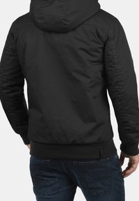 Black, hooded jacket with ribbed cuffs and hem. Features a smooth, matte finish and a relaxed fit, paired with dark blue jeans.
