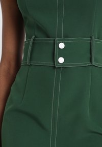 Robe vert foncé avec des coutures blanches, dotée d'une taille cintrée et d'une large ceinture ornée de deux boutons ronds en argent. Texture de tissu lisse, design taillé.