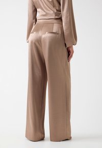 Frau trägt locker sitzende, hoch taillierte, beigefarbene Satin-Hose und passendes langärmliges Oberteil, steht vor einem schlichten weißen Hintergrund.