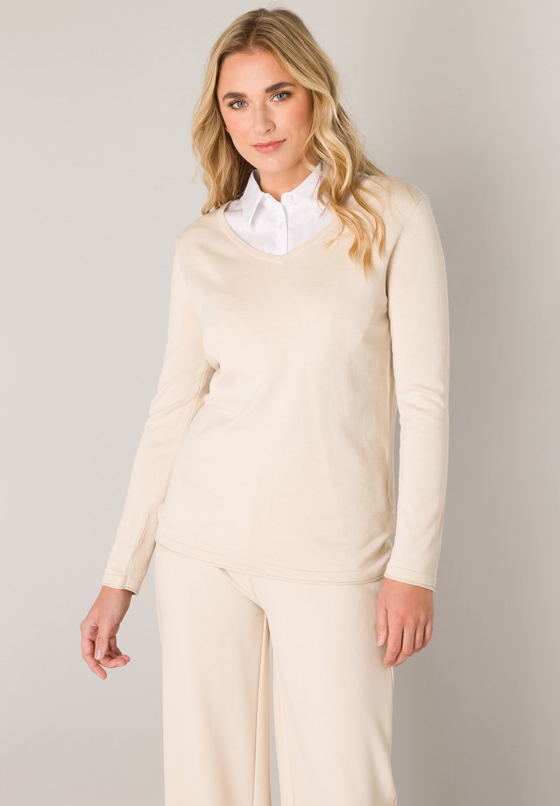Beige lange mouwen V-hals top gemaakt van zachte stof, met een gladde textuur en een ontspannen pasvorm, gedragen over een witte blouse met kraag.