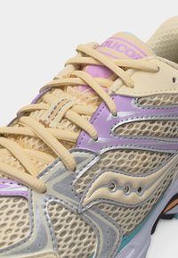 Κοντινό πλάνο αθλητικού παπουτσιού με δίχτυ σε μπεζ, λεβαντί και ασημί, με μπεζ κορδόνια και γλώσσα με το λογότυπο "SAUCONY".