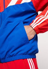 Blå och röd vindjacka med dragkedja, med en hand i en sidoficka, elastiska ärmslut och vita randiga detaljer.