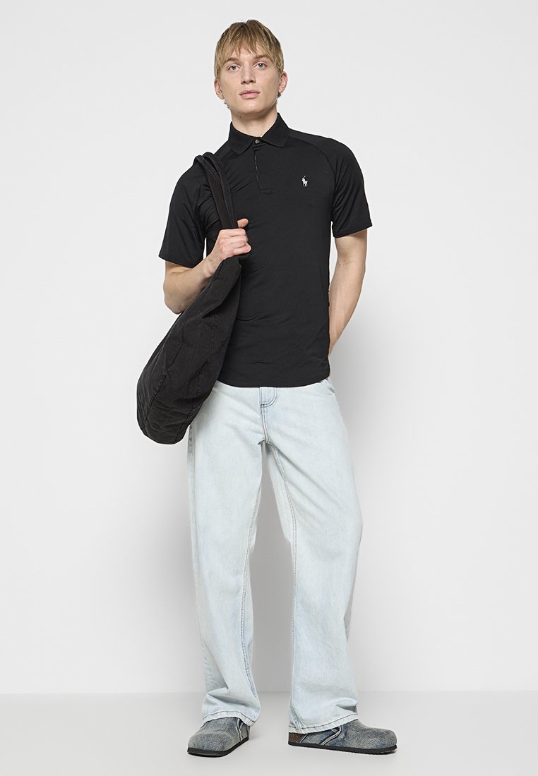 Schwarzes Poloshirt, hellblaue Jeans, graue Schuhe und eine schwarze Tasche. Das Poloshirt hat ein dezentes Logo auf der linken Brust.