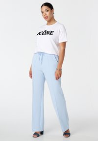 Witte t-shirt met zwart pailletten tekst "ICÔNE," gecombineerd met lichtblauwe wijde broek met een trekkoord in de taille en zijnaad.