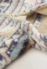 Blød fløjlsagtig babyjacket med blå, beige og cremefarvede mønstre, der har en delvist åben lynlås og synlig tøjemærke.