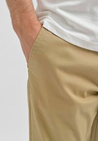 Beige bomuldsbukser med en glat tekstur, der har sidelommer og en afslappet pasform, parret med en hvid t-shirt ved taljen.